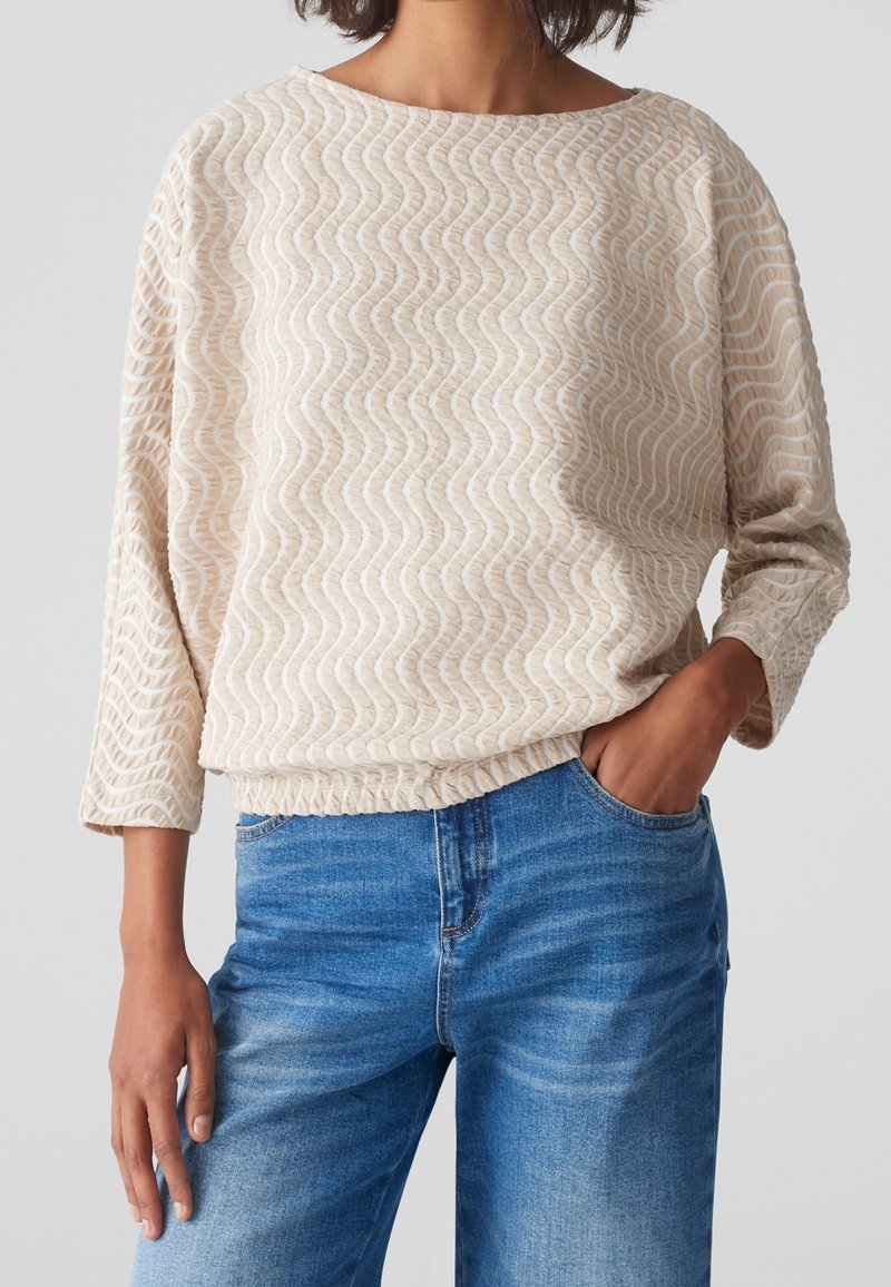 Pull beige texturé avec motif ondulé, col large et manches trois-quarts. Associé à un jean denim bleu avec une légère décoloration.