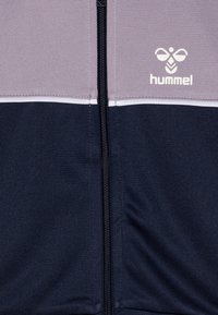 Kurtka rozpinana Hummel w kolorze szarym u góry, granatowym na dole i z białym paskiem akcentującym; wykonana z gładkiego, elastycznego materiału.