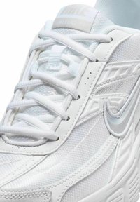Chaussure de sport blanche Nike en tissu mesh, lacets épais et surpiqûres, avec un logo Nike swoosh argenté sur le côté.