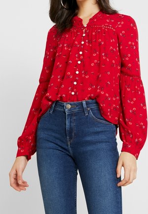 Blusa roja con flores, de mangas largas y abullonadas, con diseño de botones en la parte delantera y escote fruncido. Combinada con unos vaqueros azules de cintura alta.