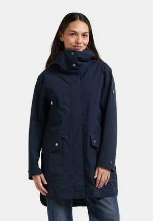 Vrouw draagt een donkerblauwe capuchonregenjas met drukknoopzakken en een losse pasvorm over een blauwe spijkerbroek, steekt zij tegen een effen achtergrond.