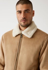 Beige Wildlederjacke mit weißem Schafsfellkragen, ausgestattet mit einem Frontreißverschluss und seitlichen Taschen, die eine glatte Textur und modernes Design präsentieren.