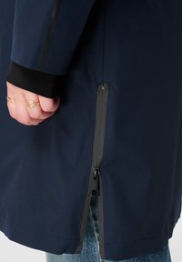 Marineblauwe waterdichte jas met een ritssluiting aan de zijkant, zwarte manchetten en een gladde textuur. Gelaagd over lichtblauwe jeans.