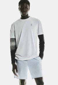 Korteband witte t-shirt met zwarte lange mouwen, met een stippenpatroon op de mouwen en een klein logo op de borst. Lichtblauwe shorts.