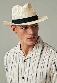 Chapeau fedora en paille avec un bandeau noir, présentant un large bord et une couronne structurée. Le modèle porte une chemise à boutons avec des rayures verticales bleues.