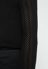 Czarny dzianinowy top z gładkim, prążkowanym korpusem i półprzezroczystym wzorem w zygzak na długich rękawach, na jednolitym tle.