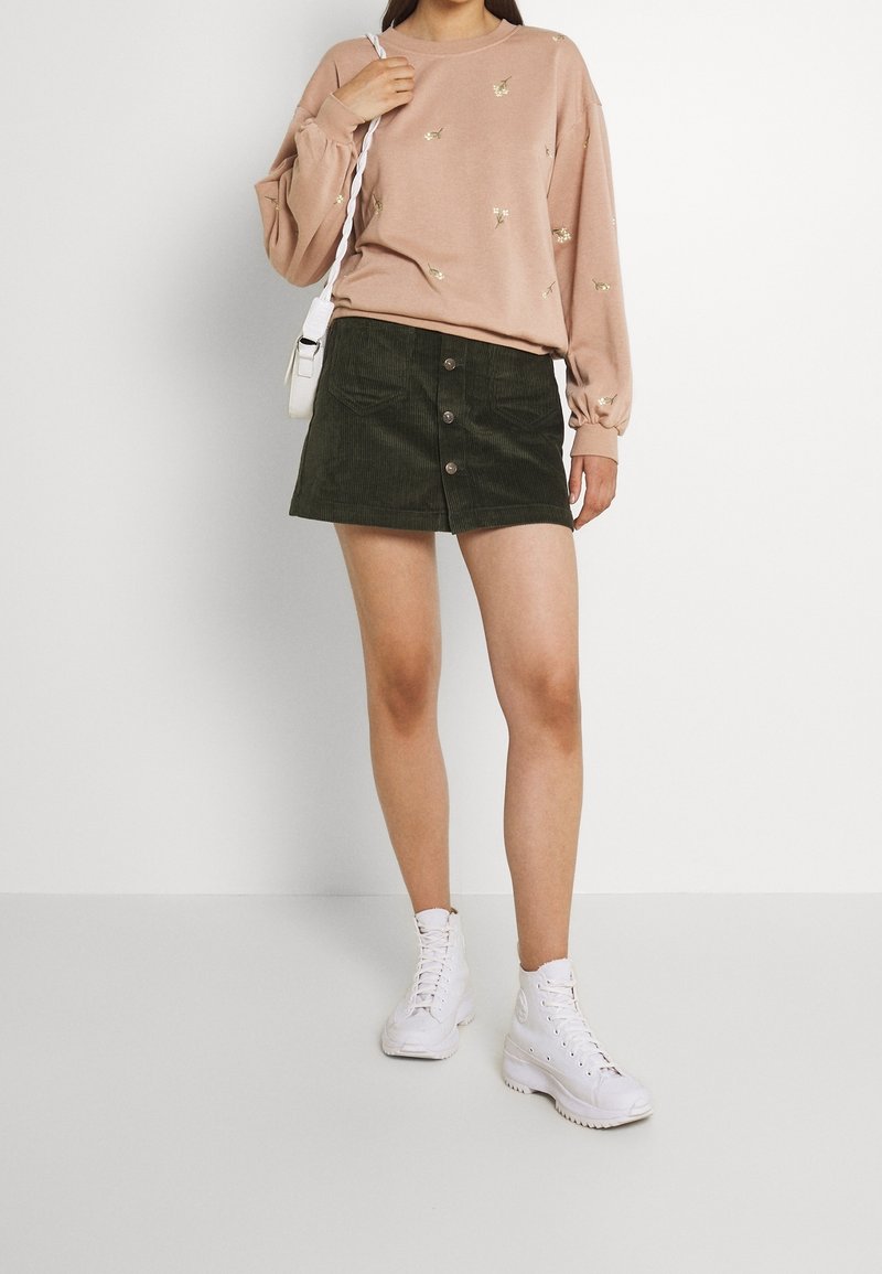 Femme portant un sweat-shirt à fleurs rose clair, une jupe en velours côtelé vert foncé boutonnée, des baskets blanches montantes, et portant un sac à bandoulière blanc.