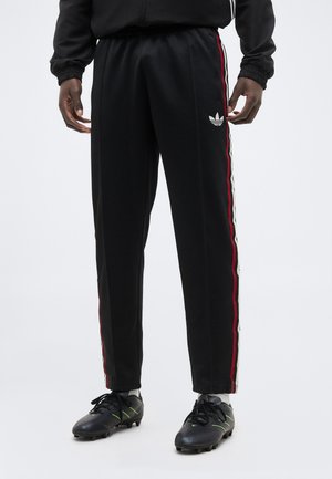 Férfi, fekete Adidas melegítőnadrágban, piros és fehér oldalsávval, fekete focicipőben, egyszínű háttér előtt állva.