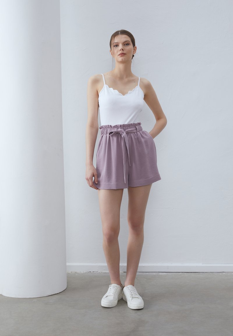 Top sin mangas blanco con bordes ondulados combinado con pantalones cortos lavanda con una cintura ajustable, hechos de tejido texturizado, y zapatillas blancas.