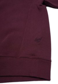 Bordeaux huppari, jossa on ribbaatut hihansuut ja helma, teksturoitu kangas ja brodeerattu logodetail alhaalla vasemmalla.