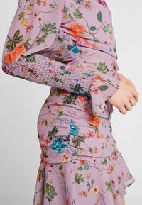 Boční pohled na osobu ve levandulových květinových šatech s nařaseným pasem a smockovými manžetami, s rukama spojenýma u pasu, na neutrálním pozadí.