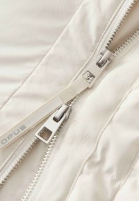 Tissu crème avec deux zippers argentés aux tirettes lisses, bords surpiqués et texture légèrement brillante. Logo visible sur la bande du zipper.