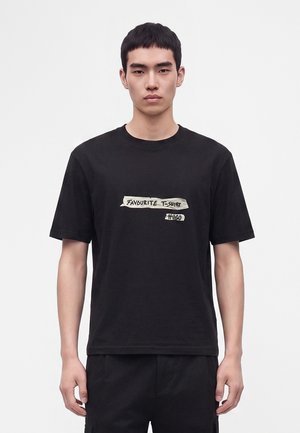 Ung mand iført en sort t-shirt med teksten "FAVOURITE T-SHIRT HUGO" i hvide bånd-lignende mærker på brystniveau.
