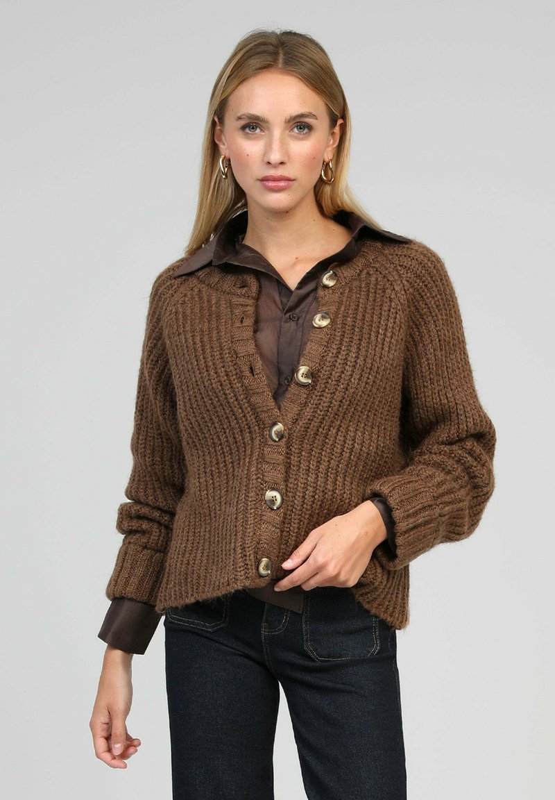 Gilet en tricot marron avec une texture côtelée, doté de grands boutons à l'avant, et d'une coupe décontractée avec des poignets roulés. Associé à une chemise sombre.
