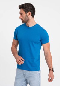 T-shirt blu in cotone con maniche corte e scollatura rotonda, indossata con jeans azzurri. Design semplice, texture liscia, forma aderente.