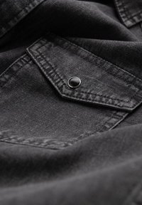 Zwarte denimstof met een gestructureerd oppervlak en zichtbare stiksels. Een kleine zwarte drukknoop accentueert het ontwerp van de puntige zak.