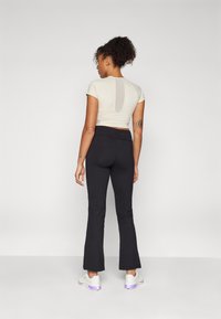 Femme aux cheveux bouclés portant un haut beige court et un pantalon noir évasé, debout de dos devant un fond uni.