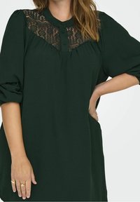 Mörkgrön blus med spetsinfällning, knappslå framtill och lösa ärmar med rynkade manschetter. Mjuk tyg med texturerade detaljer.