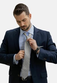 Cravate à motif diamant gris, en satin, portée avec un costume bleu marine et une chemise bleu clair, avec une montre visible au poignet.