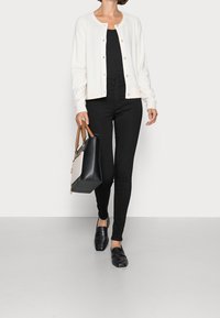 Vit cardigan med rund halsringning och knäppning, bärs över en svart topp och matchas med svarta tight jeans och loafers. Bär en svart handväska.