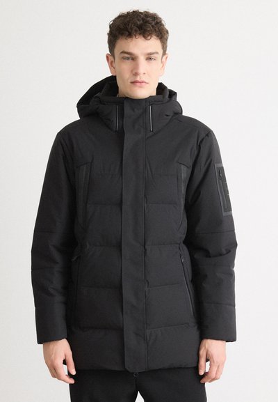 Veste noire isolée avec une capuche, présentant une texture matelassée, des poches zippées et une coupe droite. Tissu lisse avec un minimum de détails.