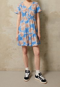 Femme portant une robe bleue à manches courtes avec un motif floral orange, des chaussettes blanches et des baskets noir et blanc, se tenant contre un mur beige.