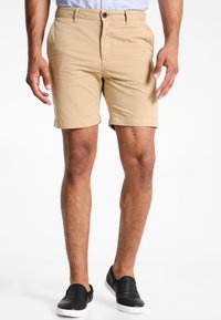 Shorts beiges en tissu léger, avec une coupe droite, des ourlets roulés et une fermeture à bouton devant. Portés avec des chaussures noires à enfiler.