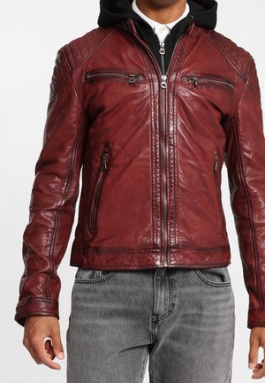 Hombre con chaqueta de cuero roja con múltiples bolsillos con cremallera y una sudadera con capucha negra debajo, combinada con jeans grises.
