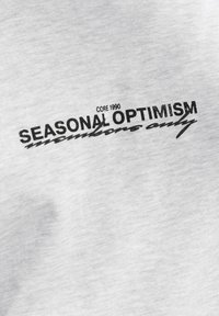 Biely textil s čiernym textom "CORE 1990 SEASONAL OPTIMISM members only" v tučnom a kurzívovom písme.