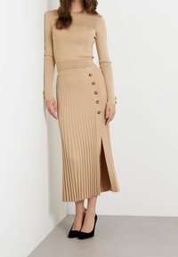 Beige pitkähihaisessa paidassa, joka on yhdistetty beigeen laskostettuun midi-hameeseen, jossa on sivusauma ja koristepainikkeet, käytettynä mustien korkokenkien kanssa.