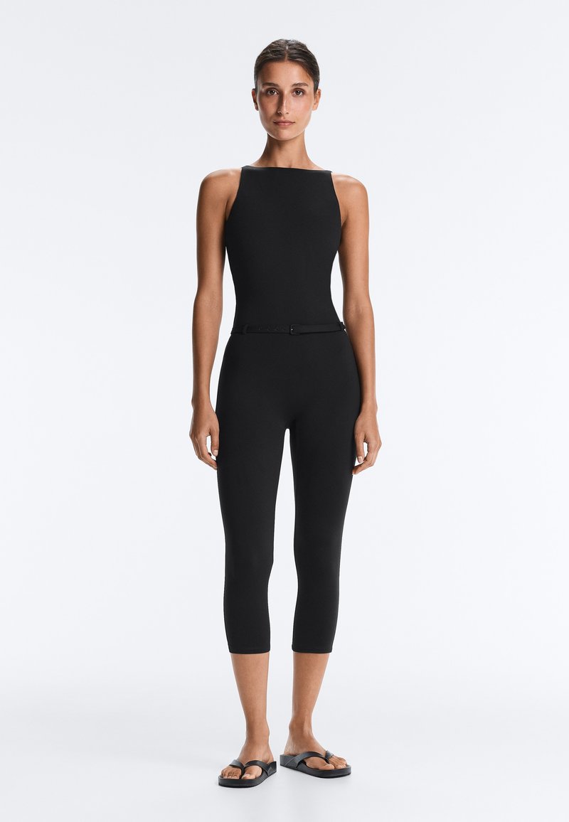 Femme debout face à l'avant portant une combinaison noire moulante sans manches avec jambes courtes et sandales plates noires sur un fond uni.