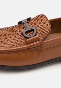 Mocassino in pelle marrone con texture intrecciata, caratterizzato da un anello a scomparsa in metallo nero e dettagli cuciti lungo i bordi. Sole in gomma piatta.