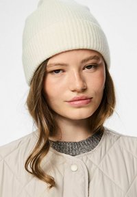 Crème ribgebreide beanie met een opgerolde rand, gedragen met een lichtbeige gewatteerde jas en een donkergrauwe trui kraag.