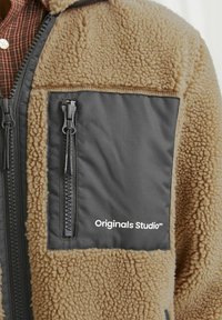 Tan fleecejacka med grå blixtlåspocketar och brodyr. Texturerad yta som liknar ull och slät nylonpanel. Inkluderar dubbelblixtlås.