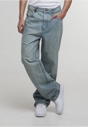 Person som har på seg løstsittende lyseblå denimjeans med hendene i lommene og hvite joggesko, står med beina i kryss mot en ensfarget bakgrunn.