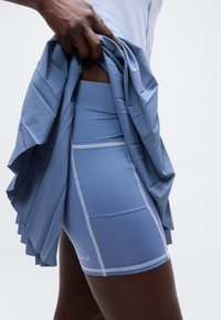 Άτομο σηκώνοντας μια γαλάζια πιετέ φούστα για να αποκαλύψει αντίστοιχο μπλε εφαρμοστό σορτς με λευκή ραφή και μικρό τυπωμένο λογότυπο στον μηρό.