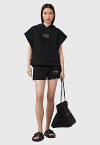 Svart oversized hoodie med en framtryckt logotyp, parat med svarta shorts med sidoknappar. Modellen håller en svart tote-väska och har på sig svarta slides.