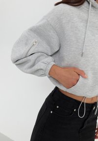 Osoba ubrana w jasnoszary krótki hoodie z zamkiem błyskawicznym na rękawie oraz czarne jeansy z wysokim stanem, z ręką schowaną w przedniej kieszeni.