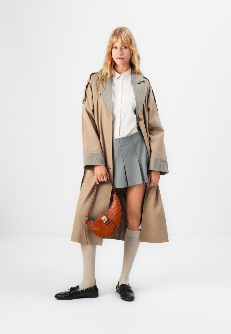 Femme portant un trench-coat beige sur une blouse blanche et une jupe plissée grise, tenant un sac à main en cuir marron, avec des chaussettes montantes et des mocassins noirs.