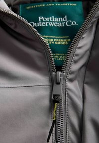 Šedá bunda s mírným leskem, se robustním zipem a zelenou nálepkou s bílým textem "Portland Outerwear Co."