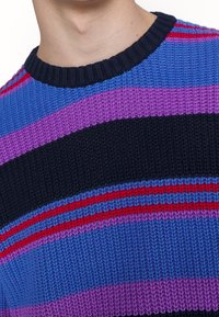 Närbild av en person som bär en stickad tröja med horisontella ränder i blå, lila, svart och röda nyanser.