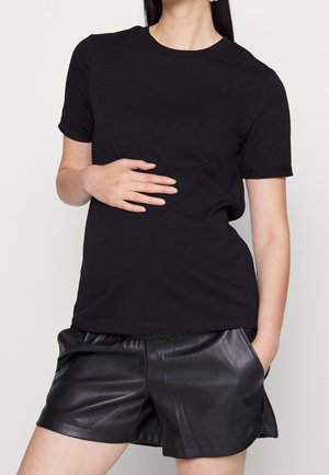 Kvinde iført sort kortærmet skjorte og sorte lædershorts, med den ene hånd på maven og den anden i lommen.