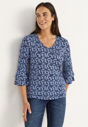 Leende kvinna klädd i en marinblå blommig topp med volanger på 3/4 ärmar och blå jeans, stående mot en vit vägg.