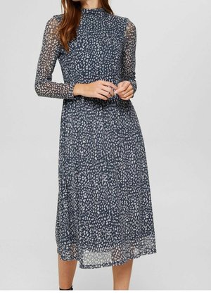 Femme portant une robe midi à manches longues bleu marine avec un motif abstrait tacheté blanc, debout devant un fond uni gris clair.