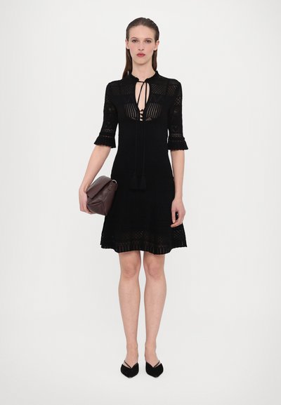 Schwarzes Strickkleid mit Spitzendetails, kurzen Ärmeln und einem Schlüsselaugen-Ausschnitt. Kombiniert mit einer braunen Clutch und schwarzen spitzen Heels.