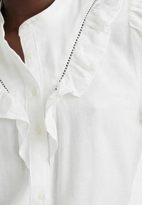 Vit skjorta med knappar i lätt tyg och med en volangkrage, med svart accentstickning längs volangens kant.