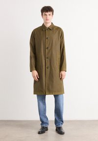 Olive green long coat with a button-down front, featuring a shirt collar and two side pockets, paired with light blue jeans and black shoes.