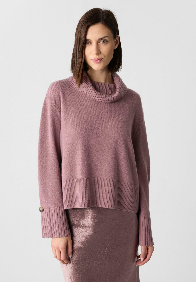 Übergroßer Pullover mit Kapuze aus weichem, pinkem Strick, mit gerippten Ärmelbündchen und Saum. Getragen mit einem glänzenden, pinken strukturierten Rock.