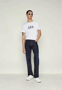 Balta kokvilnas T-kreklu ar "GAP" logo tumši zilā krāsā, apvienota ar tumši zilām džinsu biksēm. Modelis valkā baltas sporta kedas un saulesbrilles.