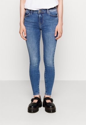 Jeans ajustados en un denim azul medio con un diseño desgastado. Presenta un estilo de cinco bolsillos, trabillas para cinturón y un cierre de botón. Combinados con zapatos negros.